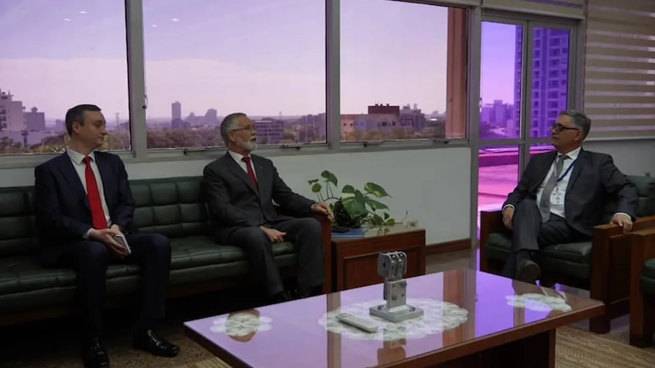 Tres hombres de traje conversan en un salón con sofás oscuros y ventanales con vista a la ciudad.