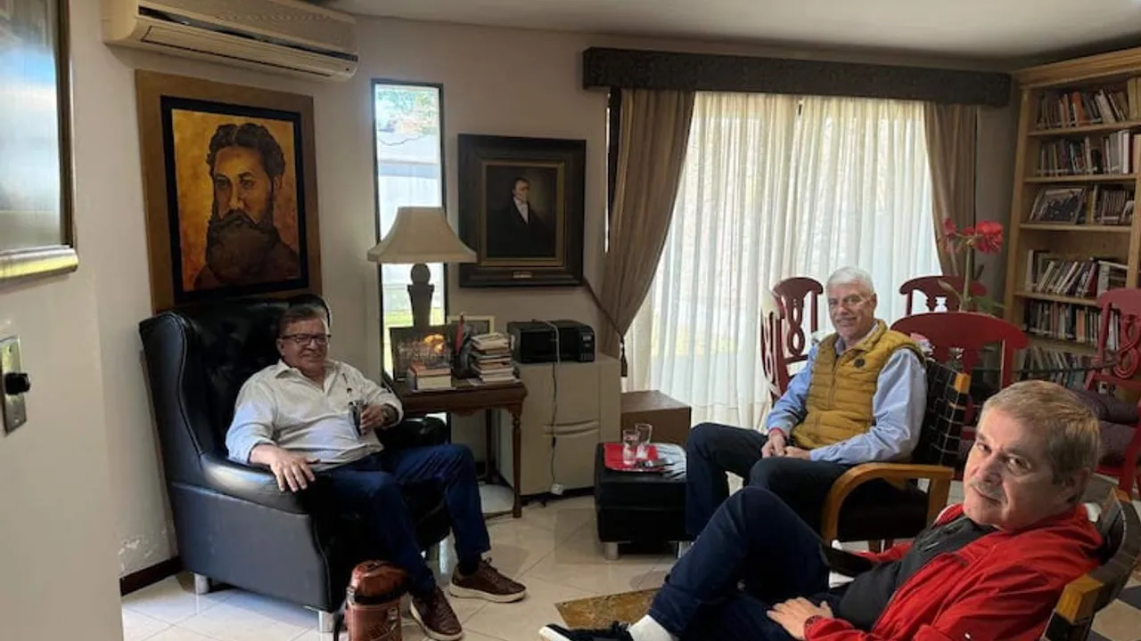 Tres hombres conversan sentados en sillones dentro de una sala con cuadros y una biblioteca.