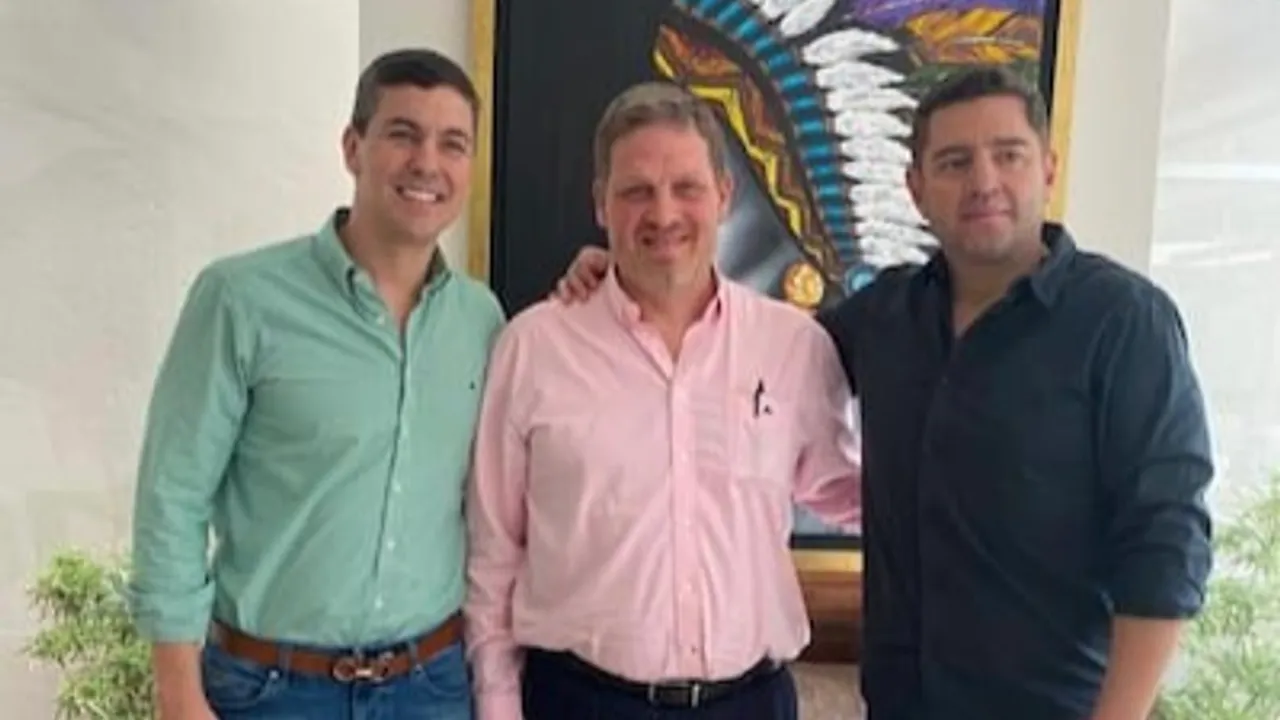 Tres hombres sonrientes parados juntos en un interior frente a una pintura decorativa.