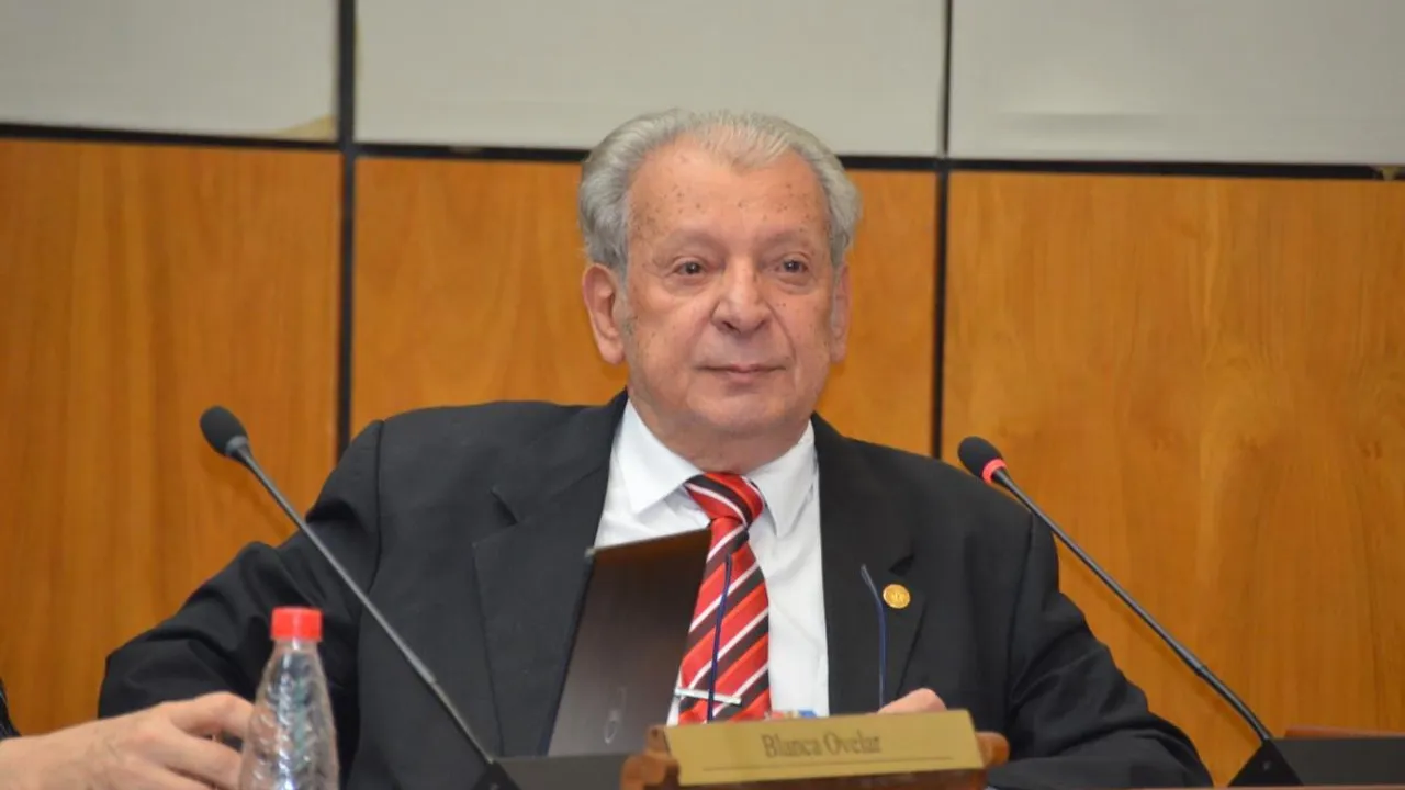 Hombre de traje oscuro y corbata a rayas sentado frente a dos micrófonos en una mesa de reuniones.