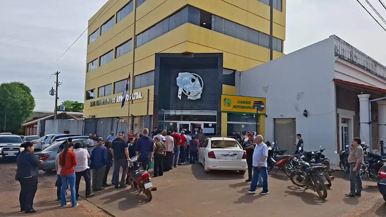Grupo de personas reunidas frente a la fachada de un edificio amarillo de la cooperativa junto a varios vehículos