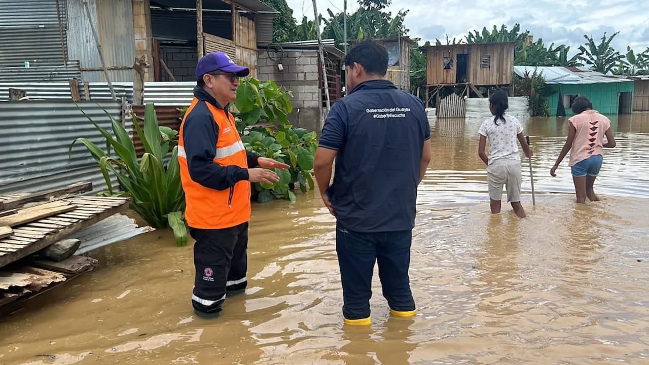 Dos funcionarios con botas de agua dialogan en una calle inundada frente a viviendas y dos personas caminando con agua al muslo.