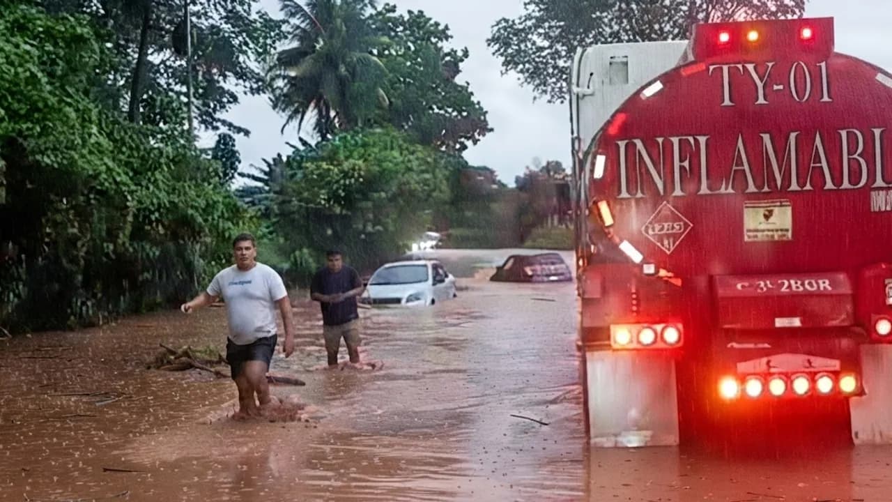 Hombres caminan con agua hasta las rodillas frente a un camión cisterna y autos varados en una carretera inundada.