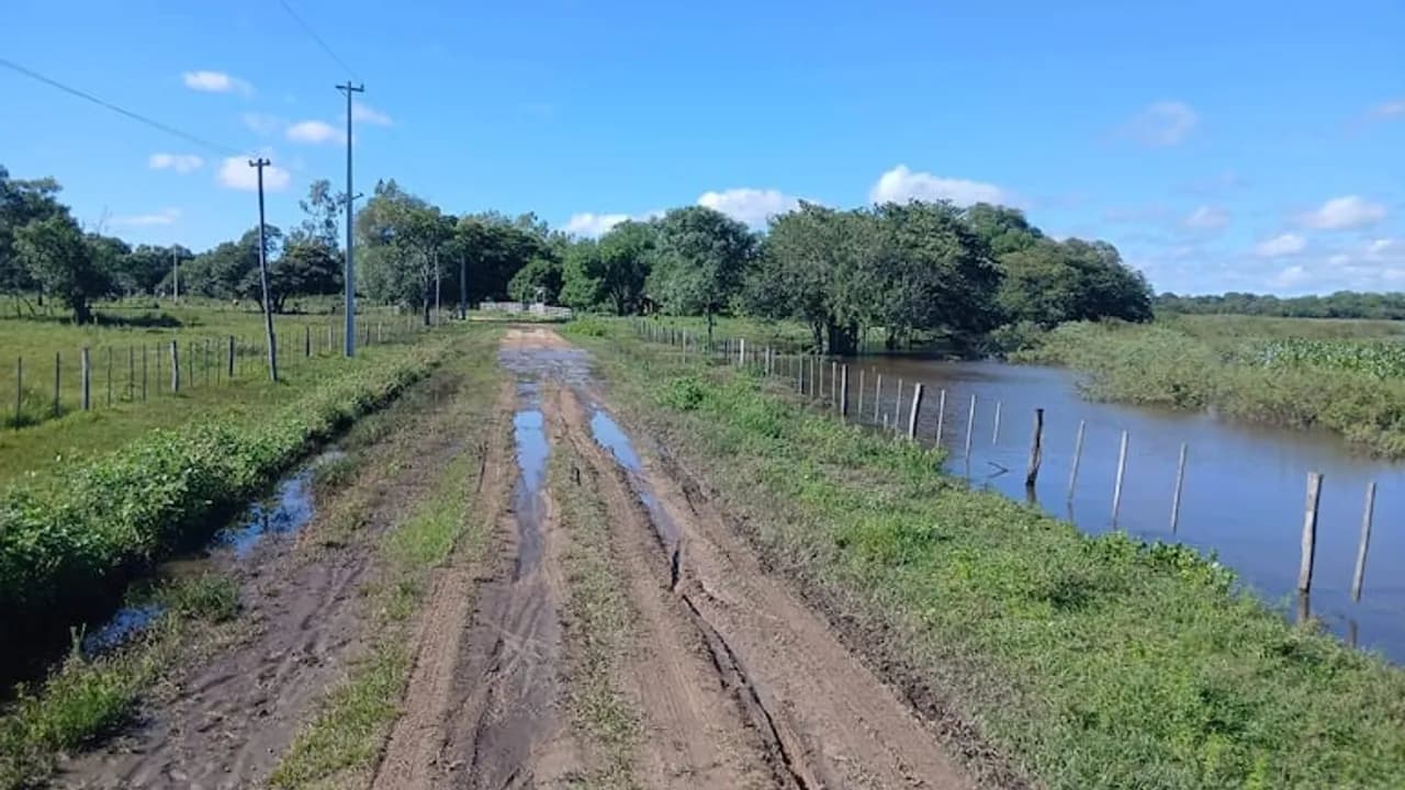 Camino rural de tierra con charcos de agua y un cauce desbordado que anega el terreno contiguo cercado.