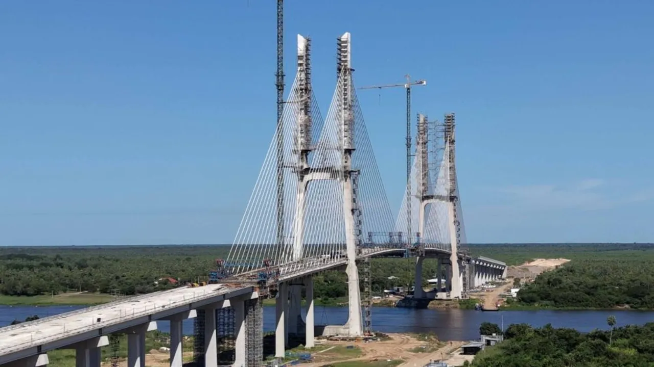 Estructura en construcción de un gran puente atirantado sobre un río rodeado de densa vegetación y cielo despejado.