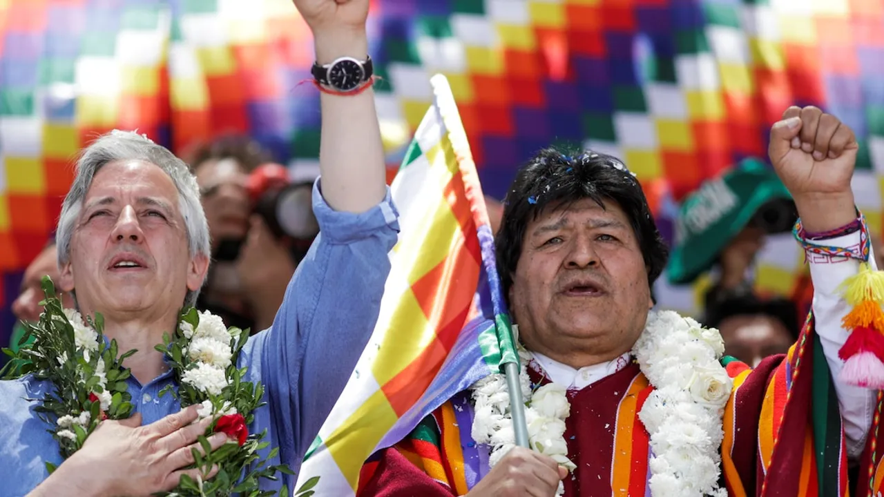 Evo Morales y Álvaro García Linera levantan el puño con guirnaldas de flores frente a una bandera wiphala.
