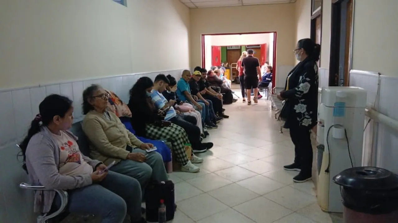 Pacientes sentados en sillas de un pasillo hospitalario esperando atención médica frente a un dispensador de agua.