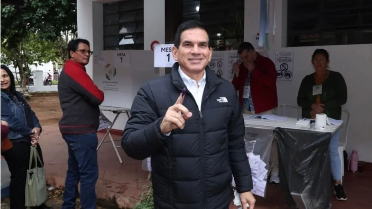 Hombre con campera oscura muestra su dedo con tinta electoral sonriendo en un local de votación.