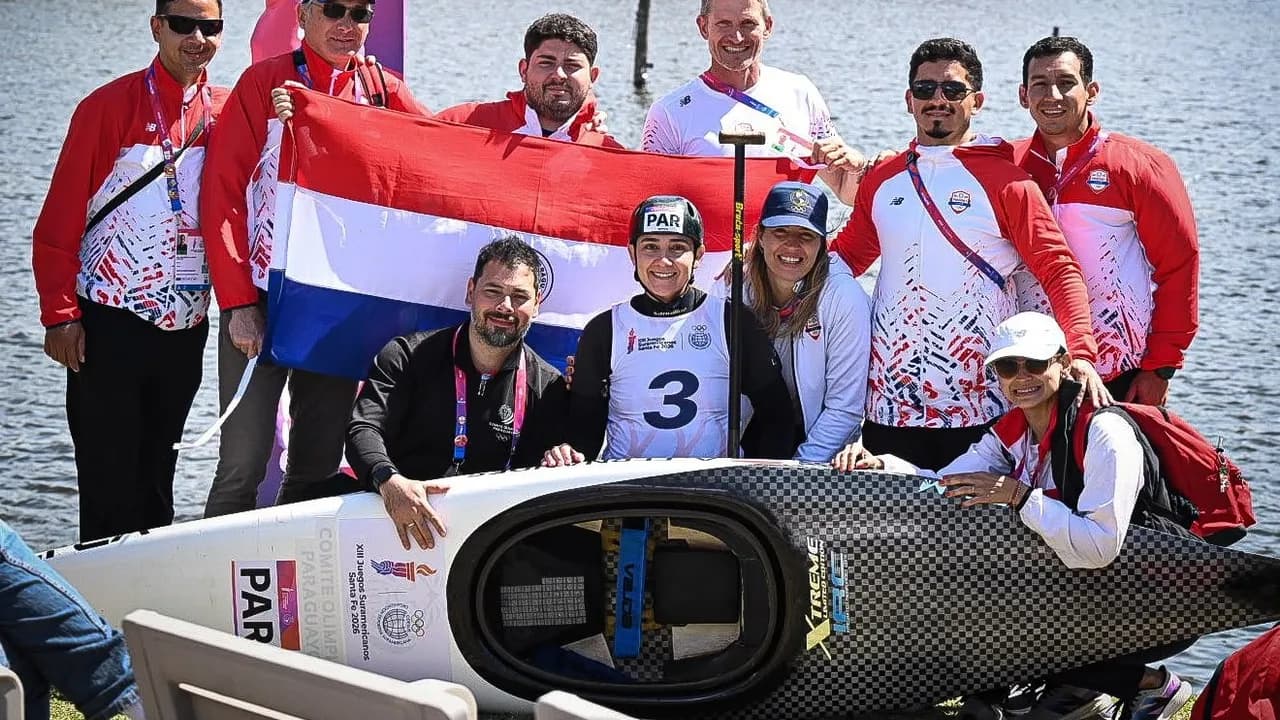 Atletas y cuerpo técnico del Comité Olímpico Paraguayo posan junto a la bandera nacional y una canoa en la orilla.