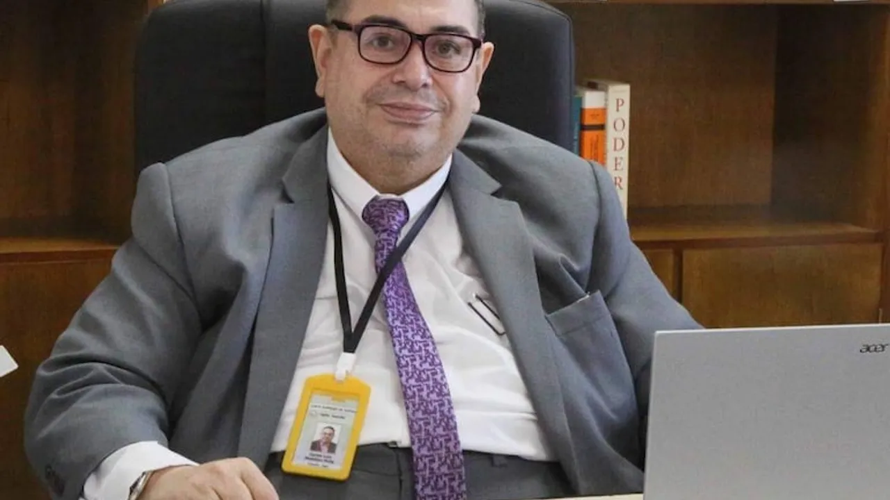 Hombre de traje gris, anteojos y corbata con diseño sentado en un escritorio de oficina frente a una computadora portátil