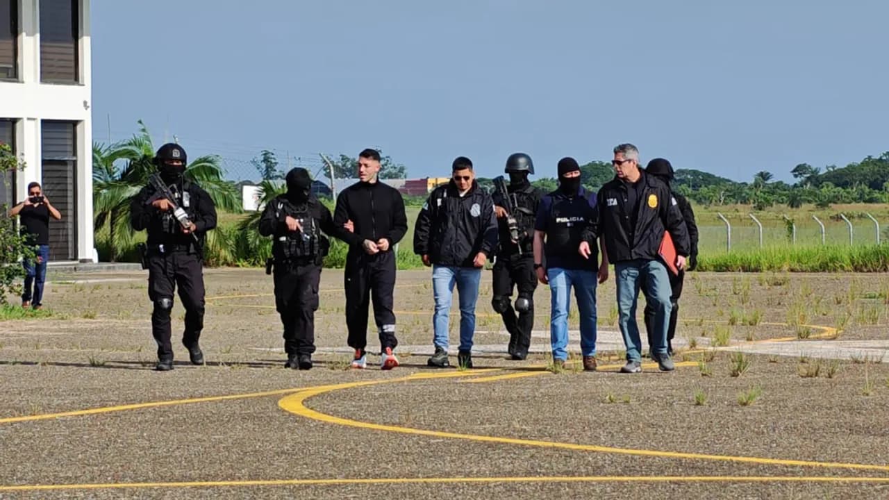 Agentes policiales y tácticos custodian a un hombre vestido de negro durante su traslado en una pista de aterrizaje.