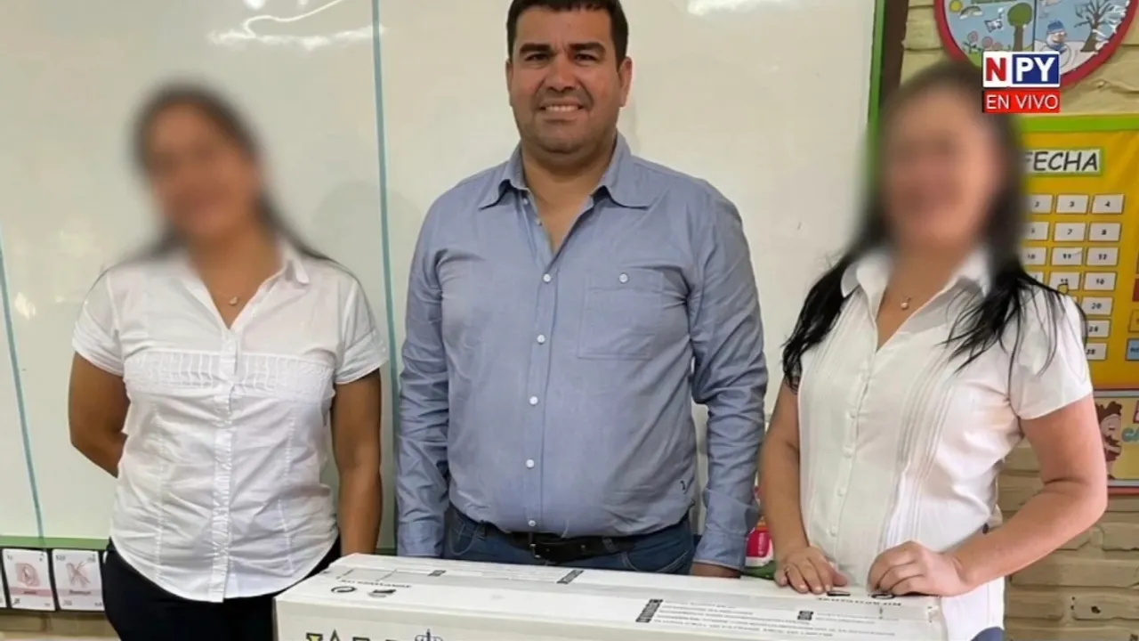 Julián Vega posa junto a dos mujeres y una caja de acondicionador de aire en un aula escolar.