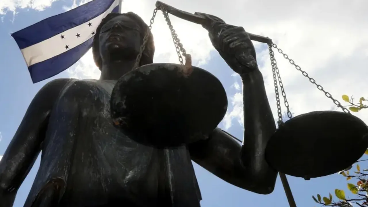 Estatua de la Dama de la Justicia con balanza en mano y la bandera de Honduras flameando de fondo bajo el cielo azul.
