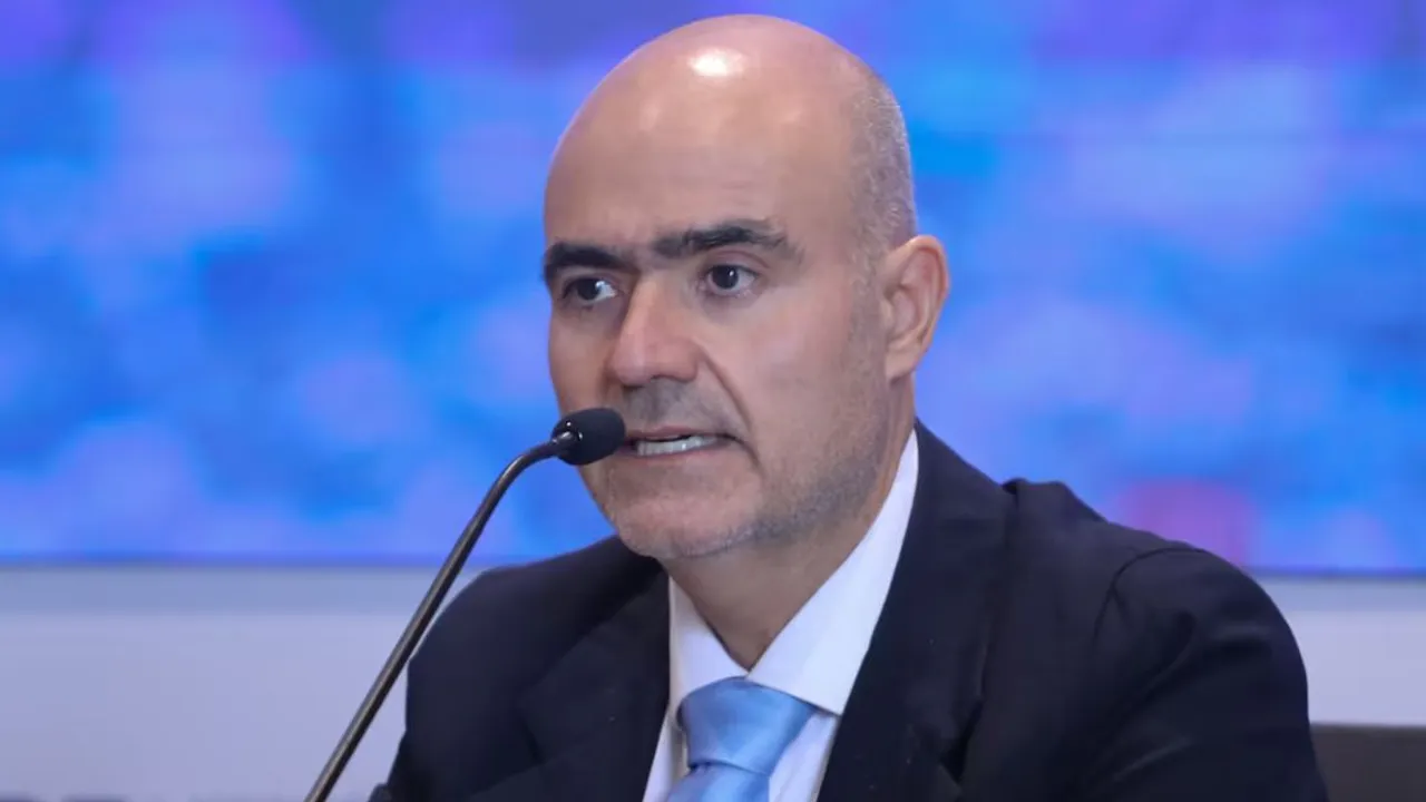 Hombre de traje oscuro, corbata celeste y camisa blanca habla frente a un micrófono en un evento institucional.