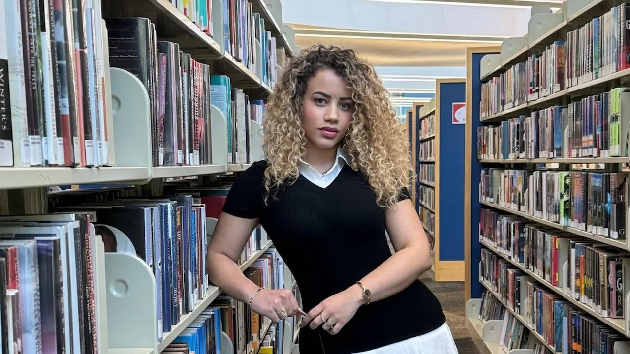 Mujer joven de cabello rubio rizado apoyada en una estantería repleta de libros dentro de una biblioteca pública.