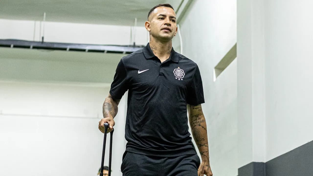 Derlis González camina por un pasillo con una remera polo negra del Club Olimpia y lleva una valija pequeña.