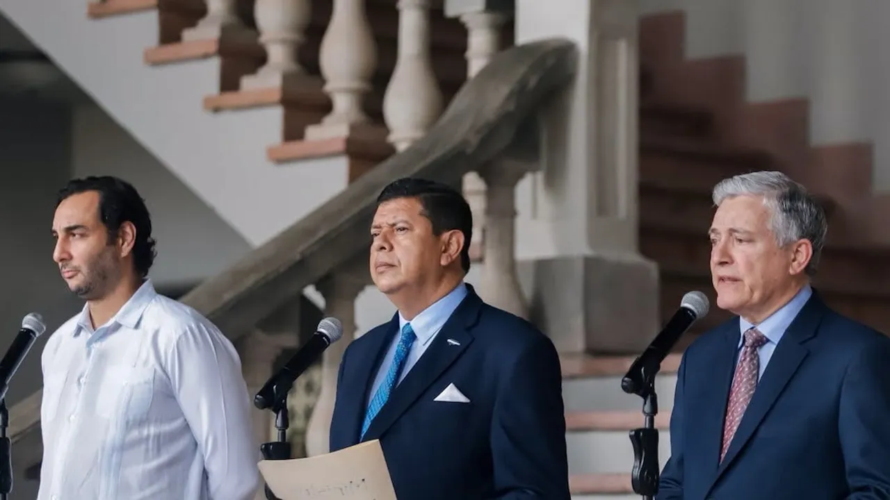 Tres funcionarios con trajes formales hablan frente a micrófonos al pie de una escalera interior.