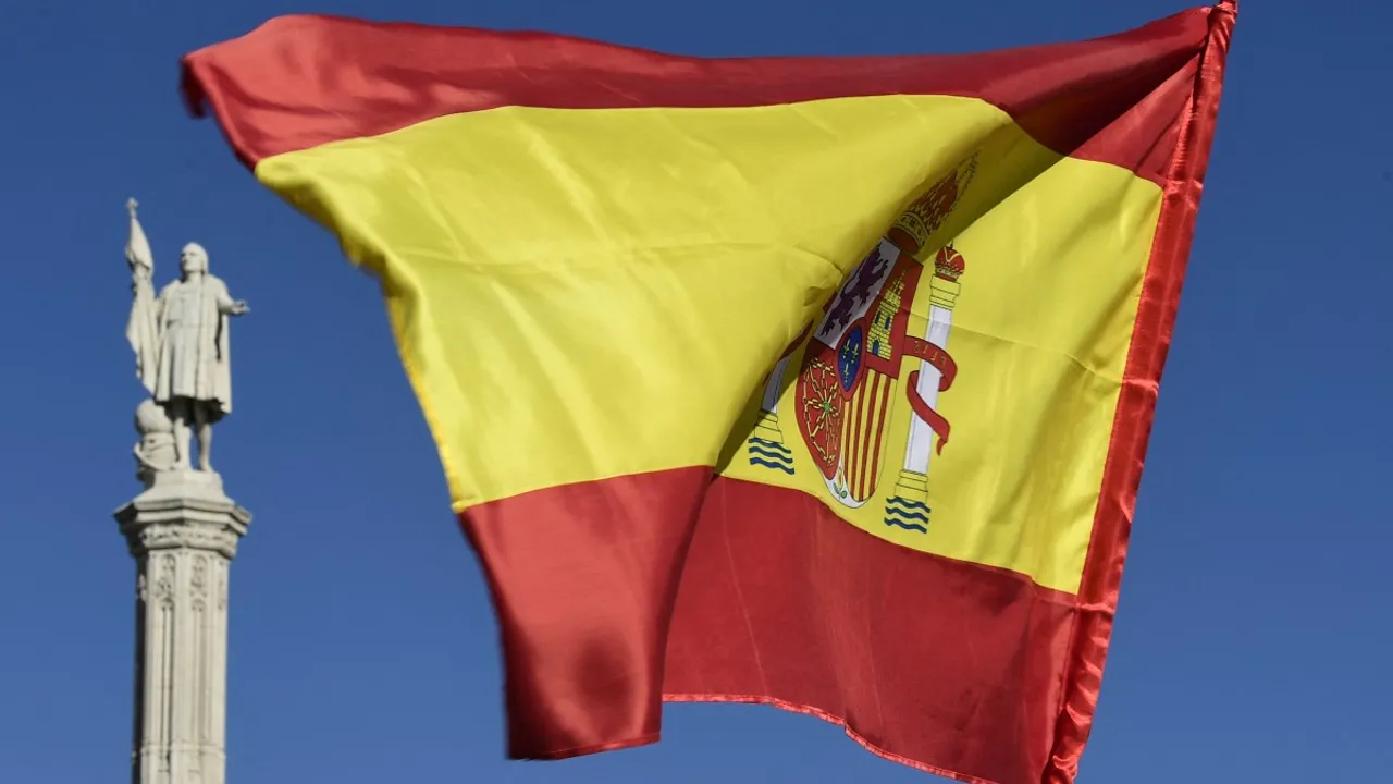 Bandera de España flameando al viento sobre un cielo azul despejado frente a un monumento histórico de piedra.