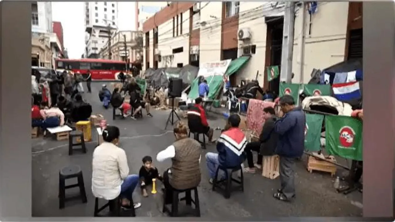 Personas sentadas en sillas sobre la calle frente a pancartas y carpas urbanas durante una manifestación