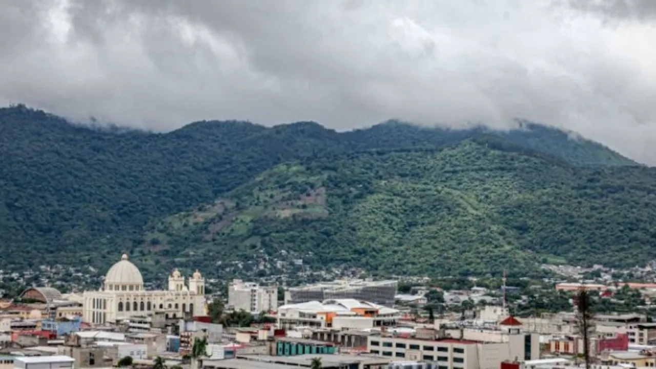 Panorámica de una ciudad con edificios y una catedral bajo montañas cubiertas por densas nubes grises.