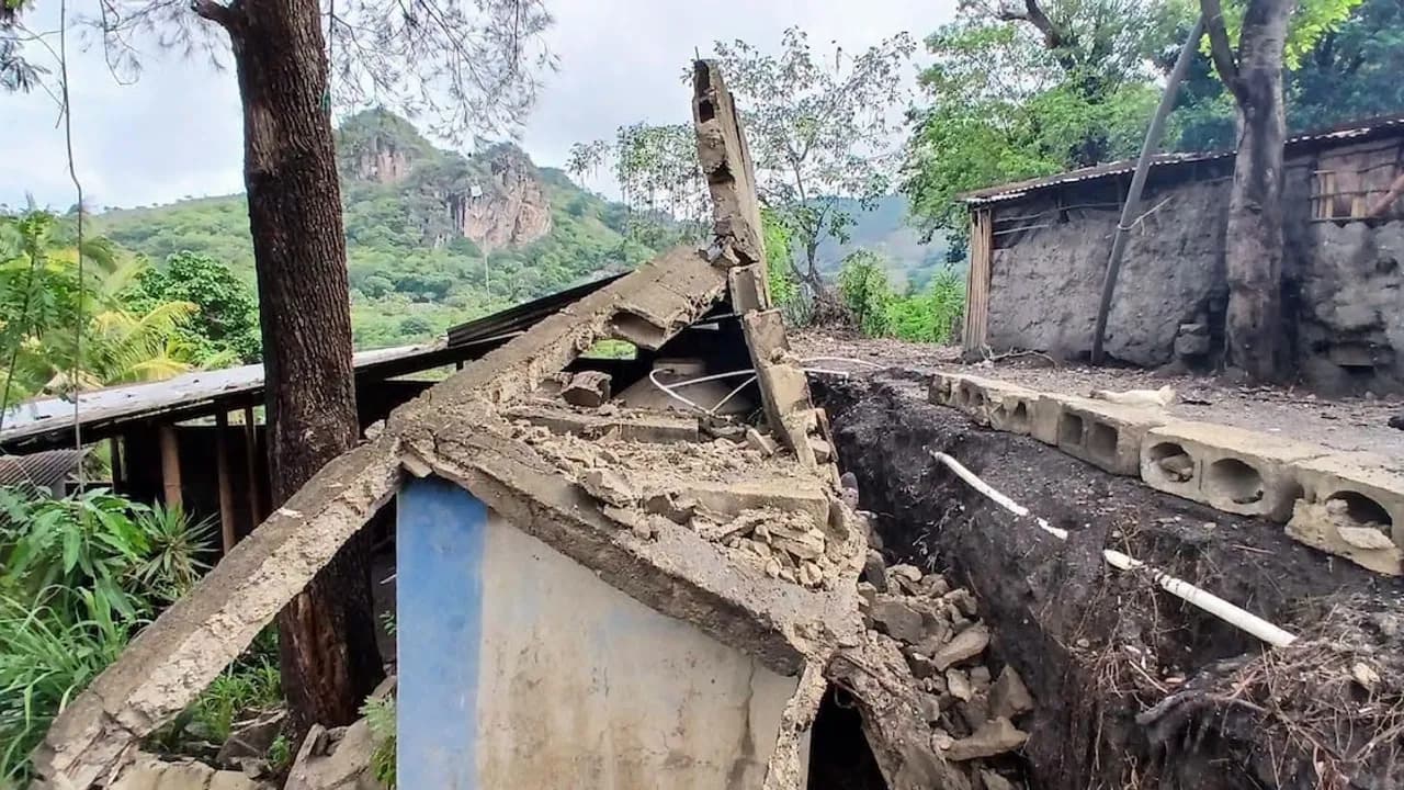 Estructura de concreto colapsada junto a una zanja de tierra con vegetación y cerros al fondo