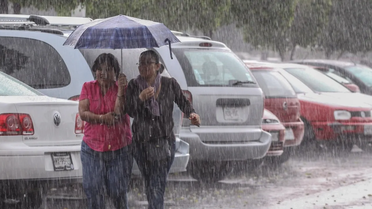 Dos mujeres caminan bajo un paraguas en medio de una fuerte lluvia en un estacionamiento de autos.