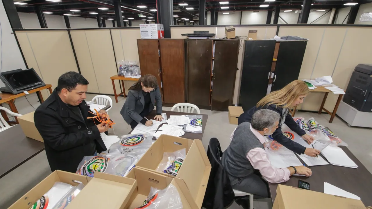 Funcionarios y apoderados sentados en mesas revisando documentos y bolsas transparentes con sellos oficiales