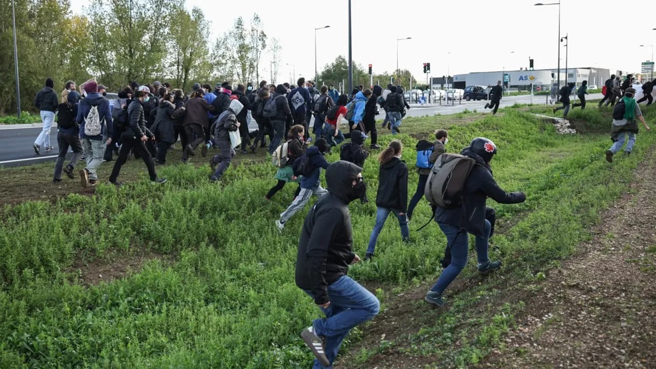 Grupo de manifestantes vestidos de negro corre por un espacio verde junto a una autopista durante una protesta.