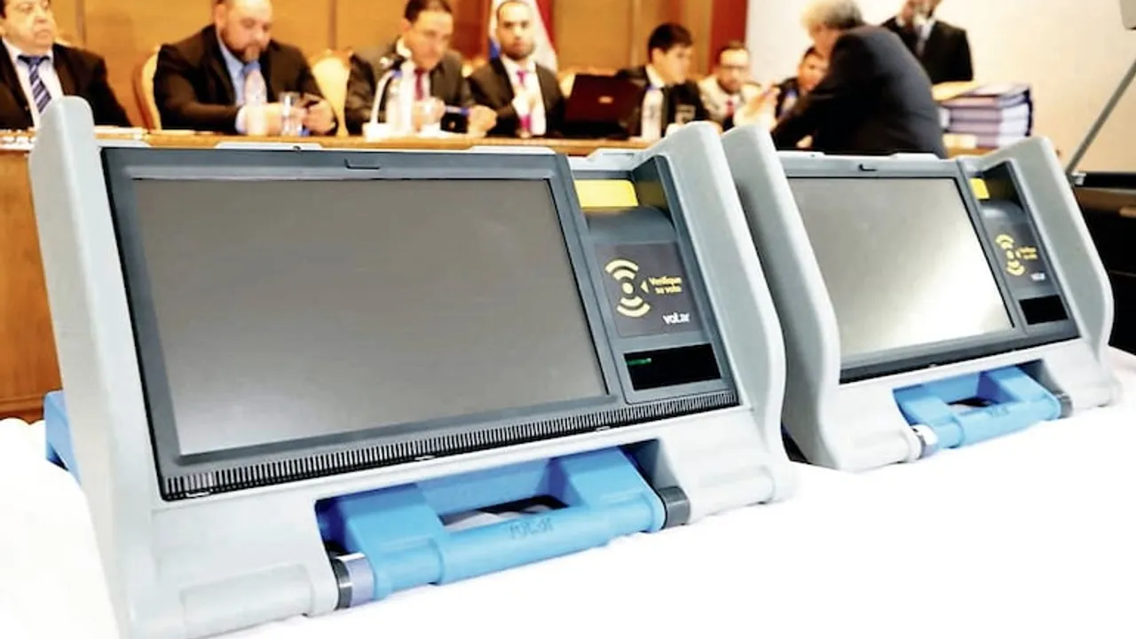 Dos urnas electrónicas con pantallas táctiles apagadas sobre una mesa ante autoridades legislativas.