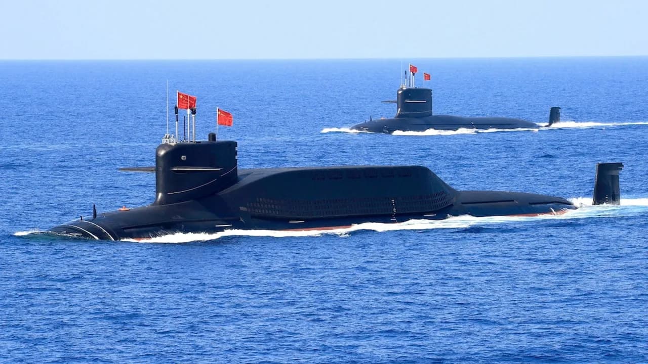 Dos submarinos militares de color negro navegan sobre la superficie del mar abierto con banderas rojas izadas.