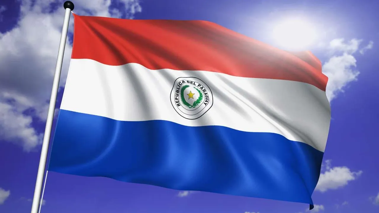 Bandera paraguaya flameando en un mástil metálico con el sol brillante y un cielo azul despejado de fondo.
