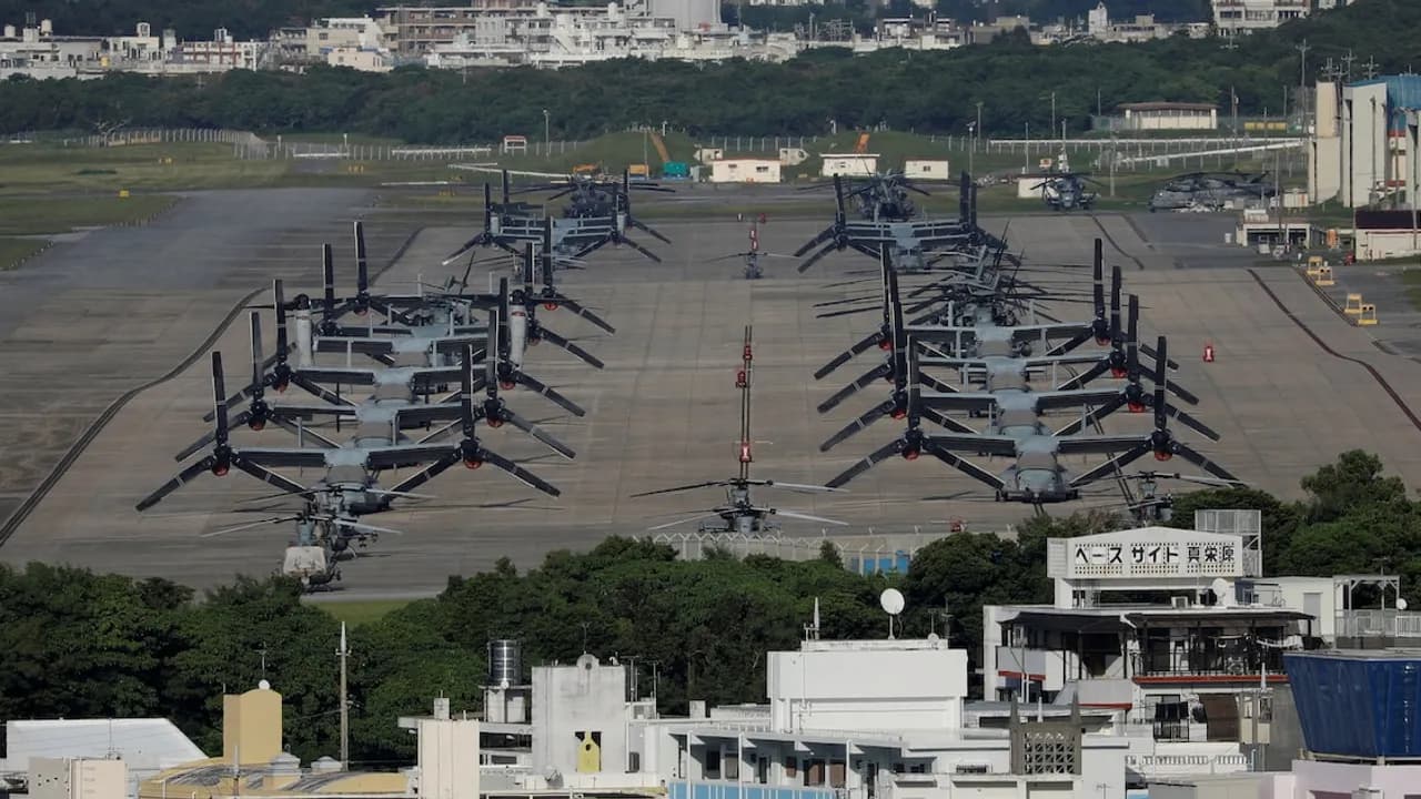 Fila de aeronaves militares MV-22 Osprey estacionadas en la pista de una base aérea con edificaciones de fondo.