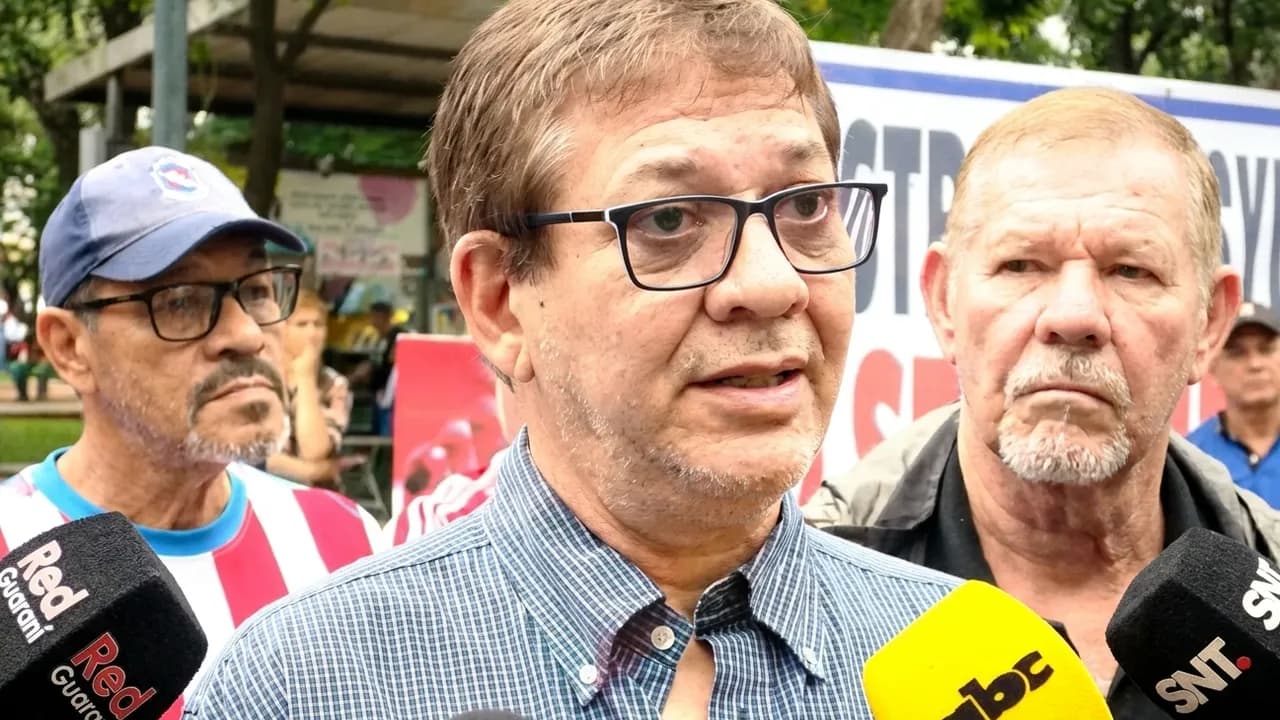 Nelson Samudio habla ante micrófonos de prensa acompañado de otros manifestantes frente a un cartel.