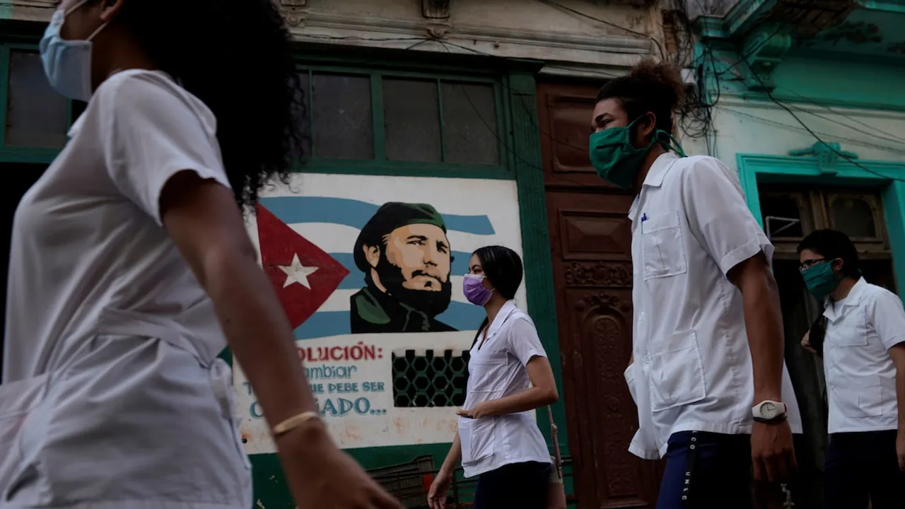 Estudiantes con batas médicas blancas y mascarillas caminan frente a un mural con la imagen del Che Guevara.