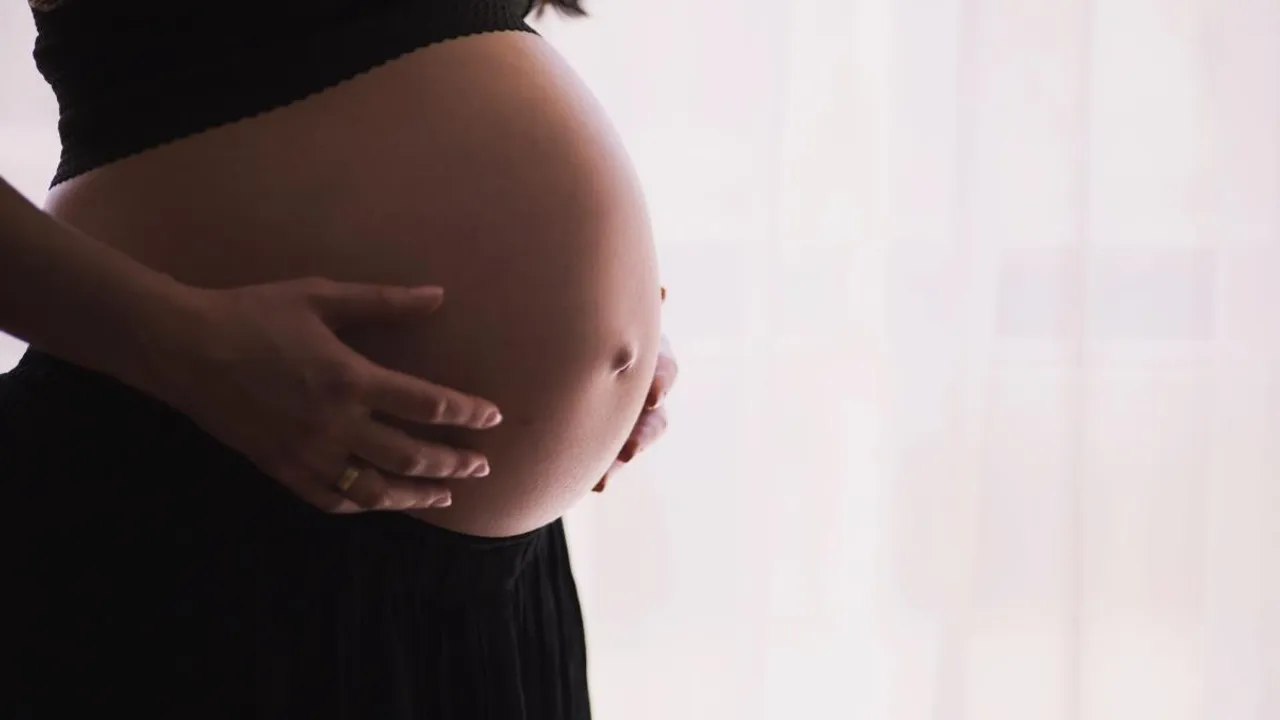 Mujer en etapa avanzada de gestación sosteniendo su vientre descubierto de perfil frente a una ventana luminosa.