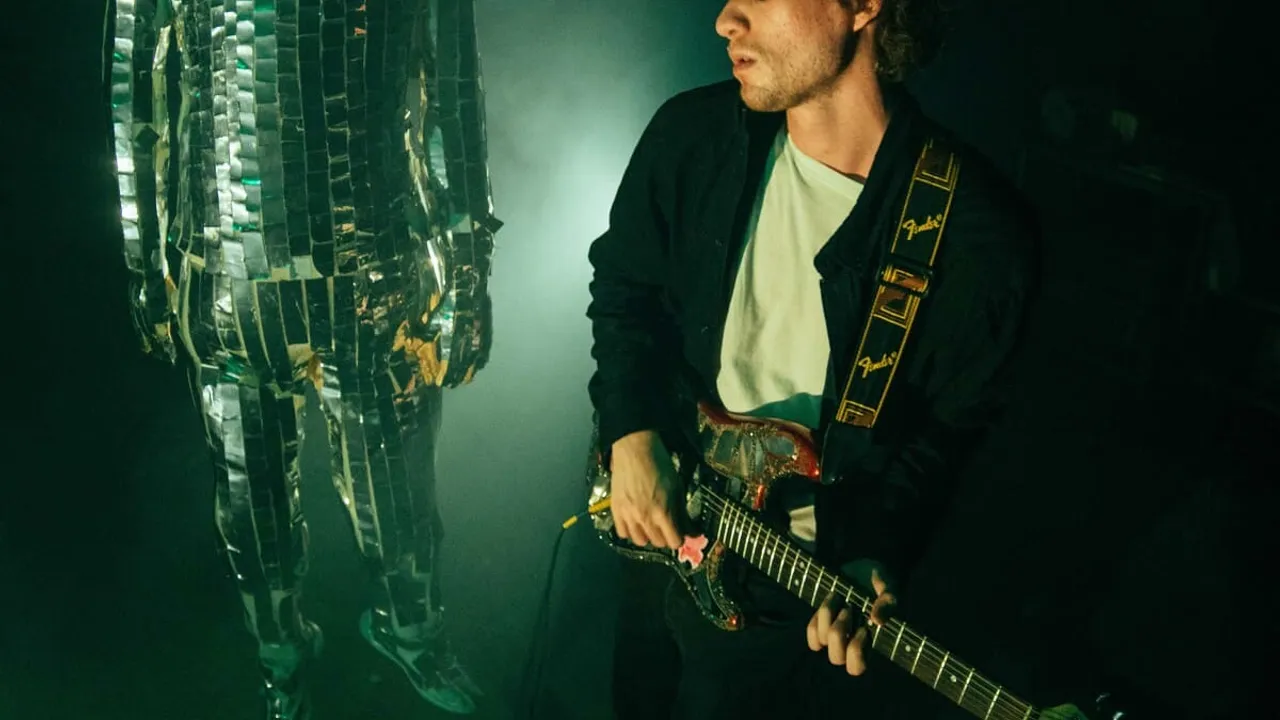 Un músico sostiene una guitarra eléctrica junto a un personaje vestido con un traje de espejos reflectantes.