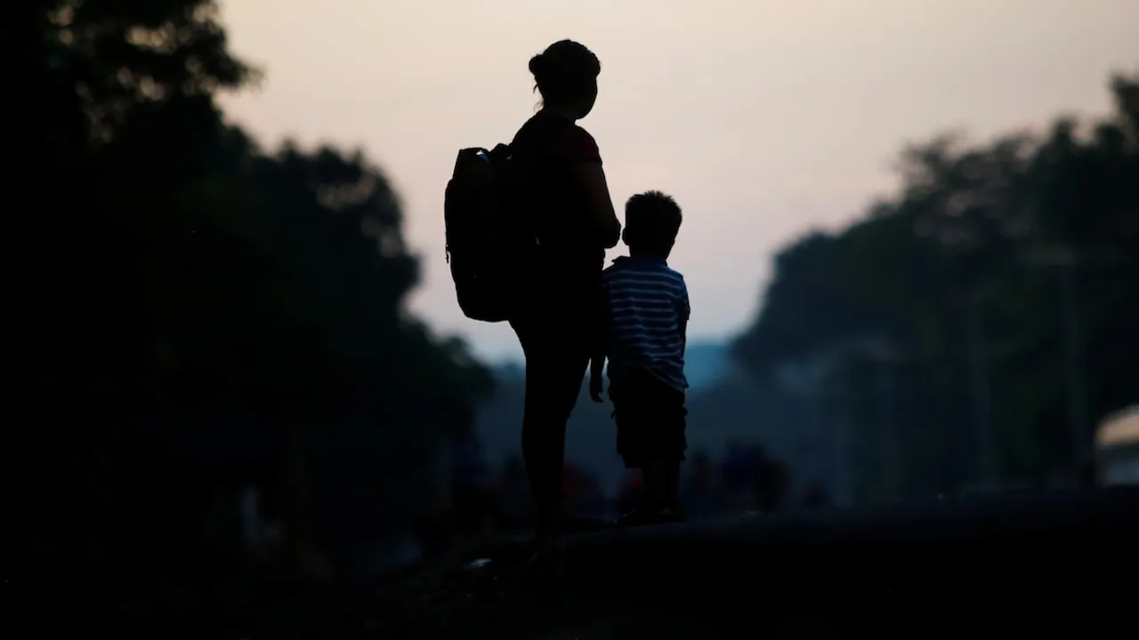 Silueta a contraluz de una mujer con mochila junto a un niño pequeño de pie al atardecer en una carretera.