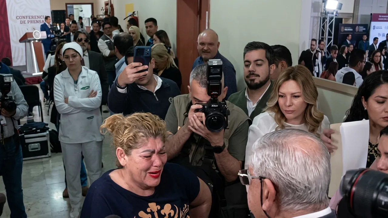 Mujer llora visiblemente conmovida mientras habla frente a una multitud y cámaras de prensa en un recinto público.