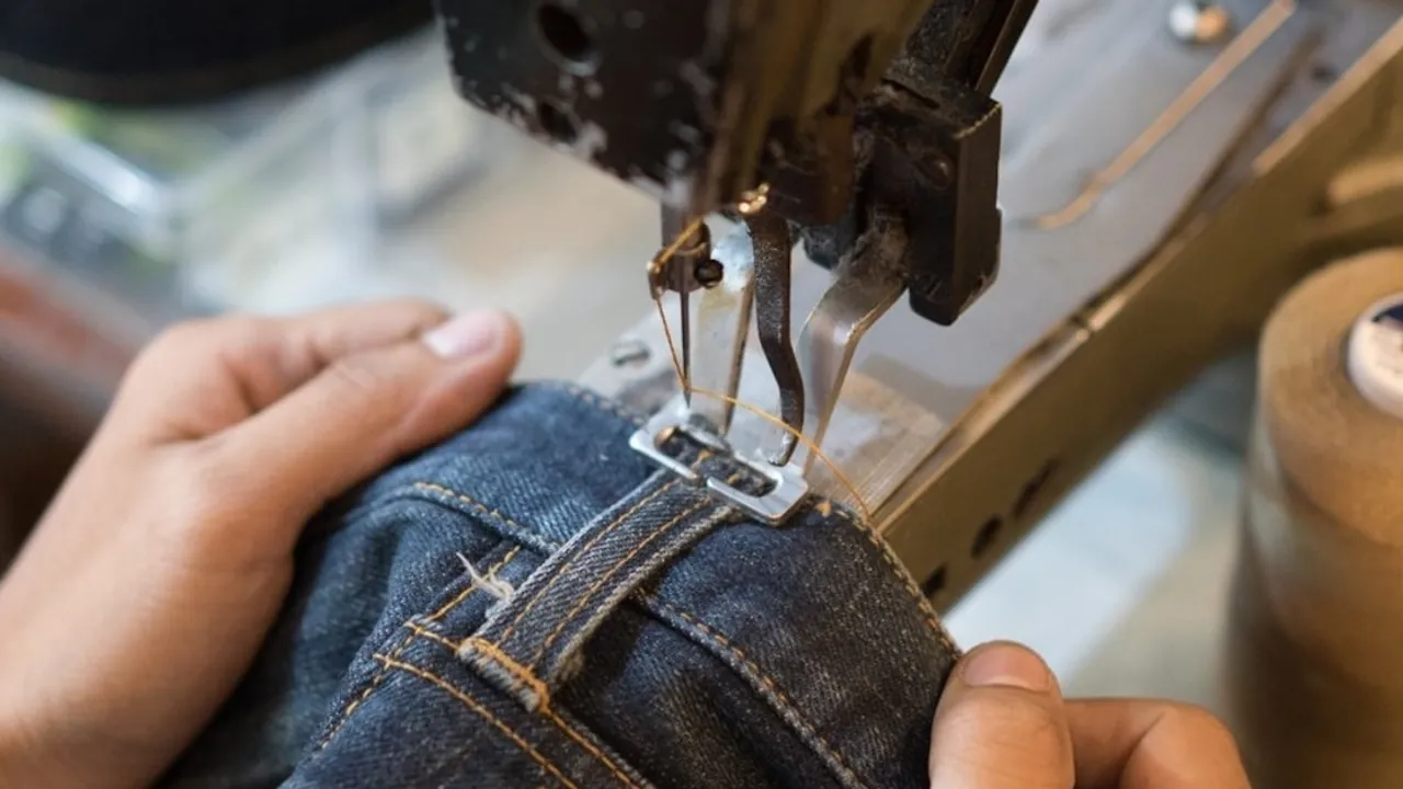 Costurero manipulando tela de mezclilla bajo la aguja de una máquina de coser industrial en un taller textil.