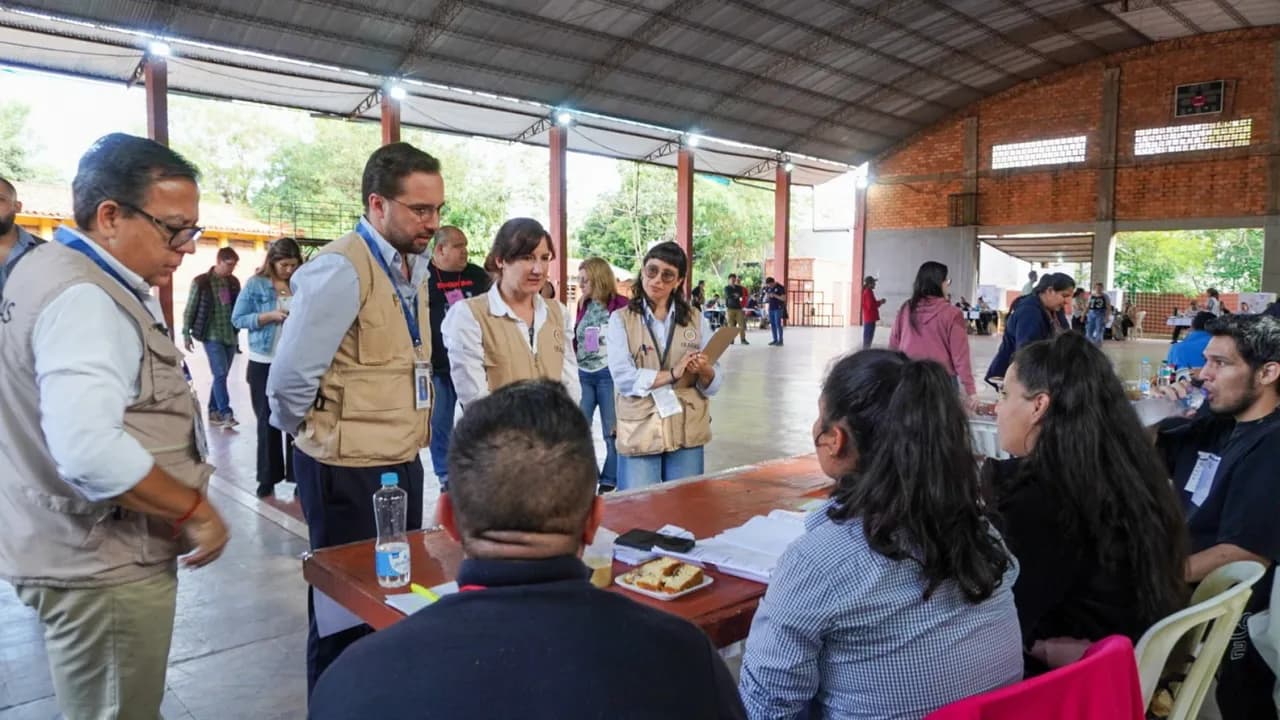 Observadores de la misión electoral de la OEA con chalecos distintivos conversan con miembros de una mesa de votación