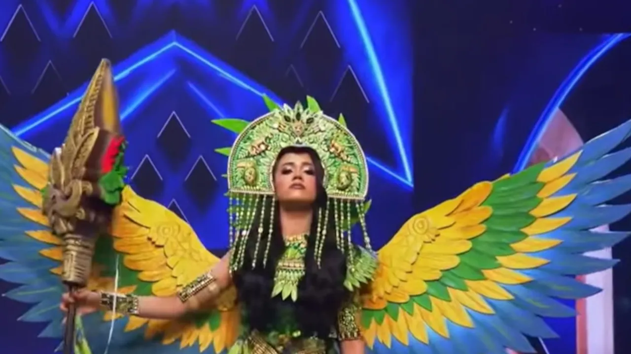 Valeria Amaya luce un atuendo con grandes alas de plumas verdes, amarillas y azules, sosteniendo un cetro ceremonial.