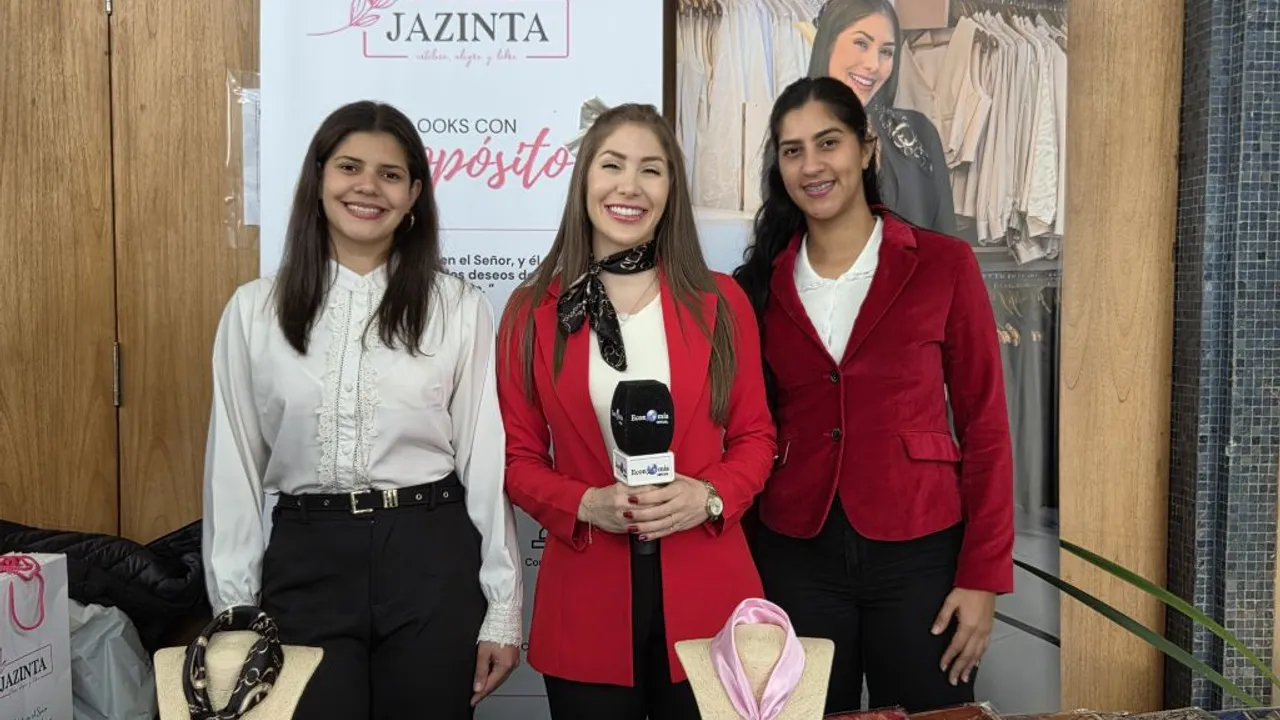 Tres mujeres vestidas con trajes elegantes posan frente a un cartel de la marca Jazinta en un evento corporativo.
