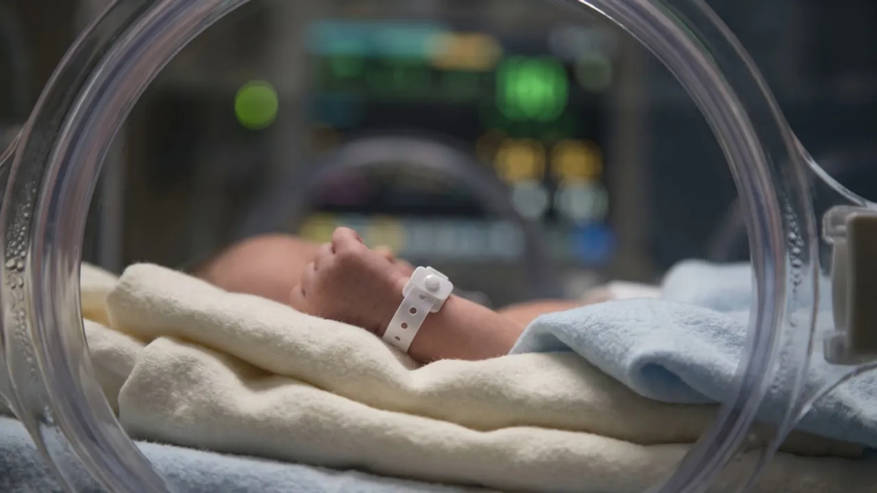 Recién nacido con pulsera de identificación médica descansa dentro de una incubadora neonatal transparente.