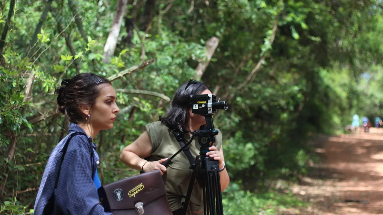 Dos mujeres manejan una cámara analógica Super 8 montada sobre un trípode en un sendero rodeado de árboles.