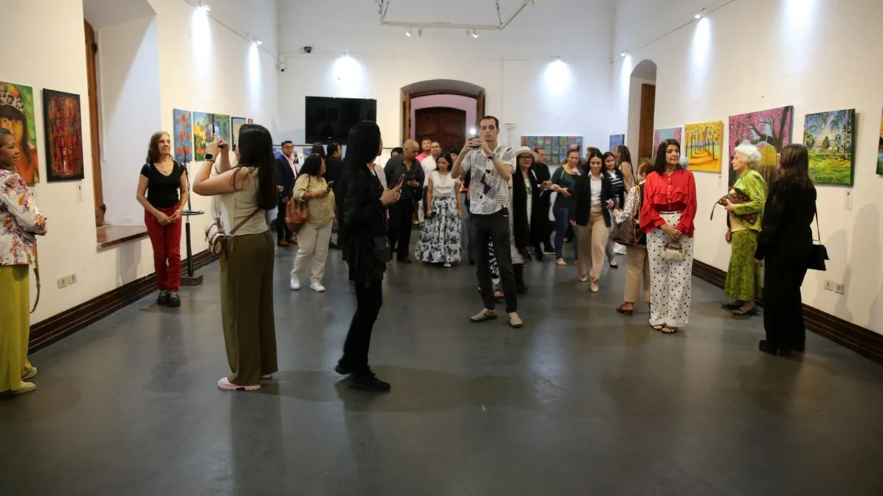 Personas recorren una sala iluminada de exposición con cuadros en las paredes y toman fotografías con sus teléfonos.