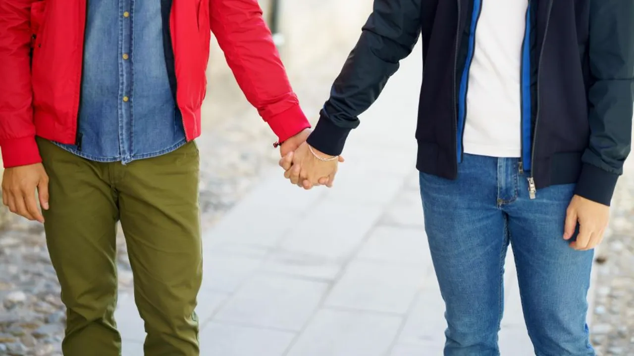 Dos personas caminan tomadas de la mano vistiendo abrigos y pantalones de tela sobre una vereda.