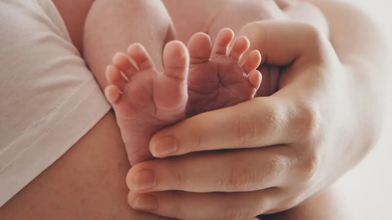 Manos adultas sostienen con delicadeza los pequeños pies descalzos de un recién nacido acostado.