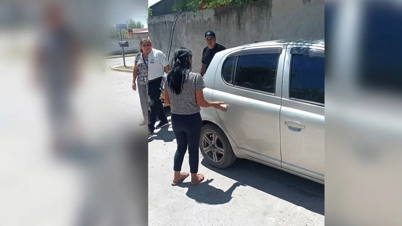 Una mujer y dos personas se acercan a la puerta trasera de un automóvil plateado en una calle soleada.