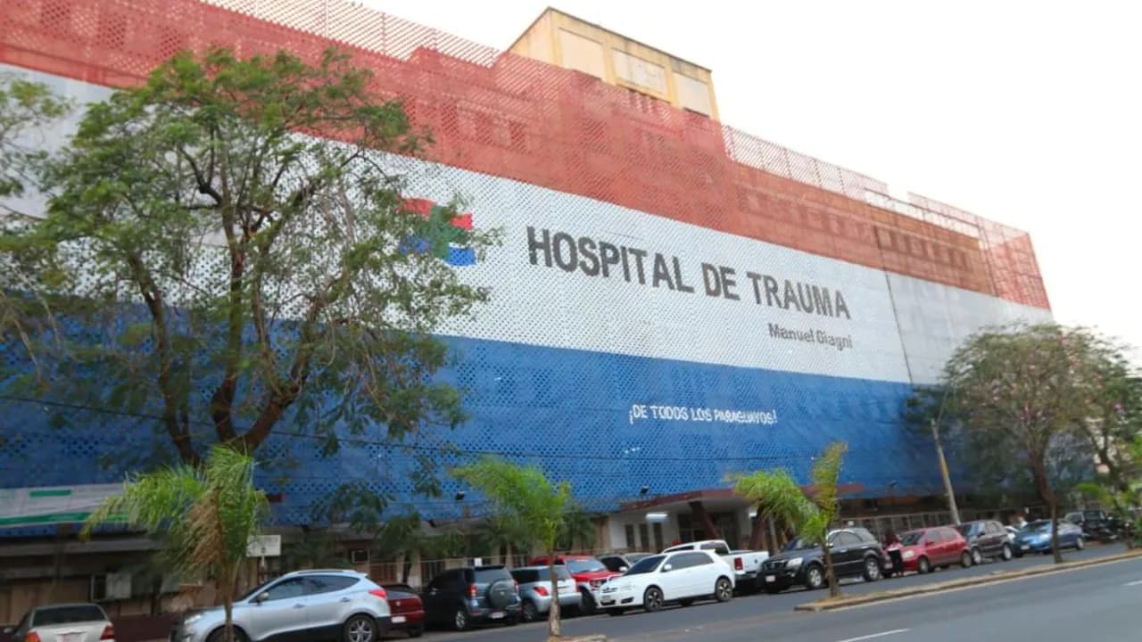 Fachada del Hospital de Trauma con red tricolor paraguaya, cartel institucional y vehículos estacionados frente al edificio.