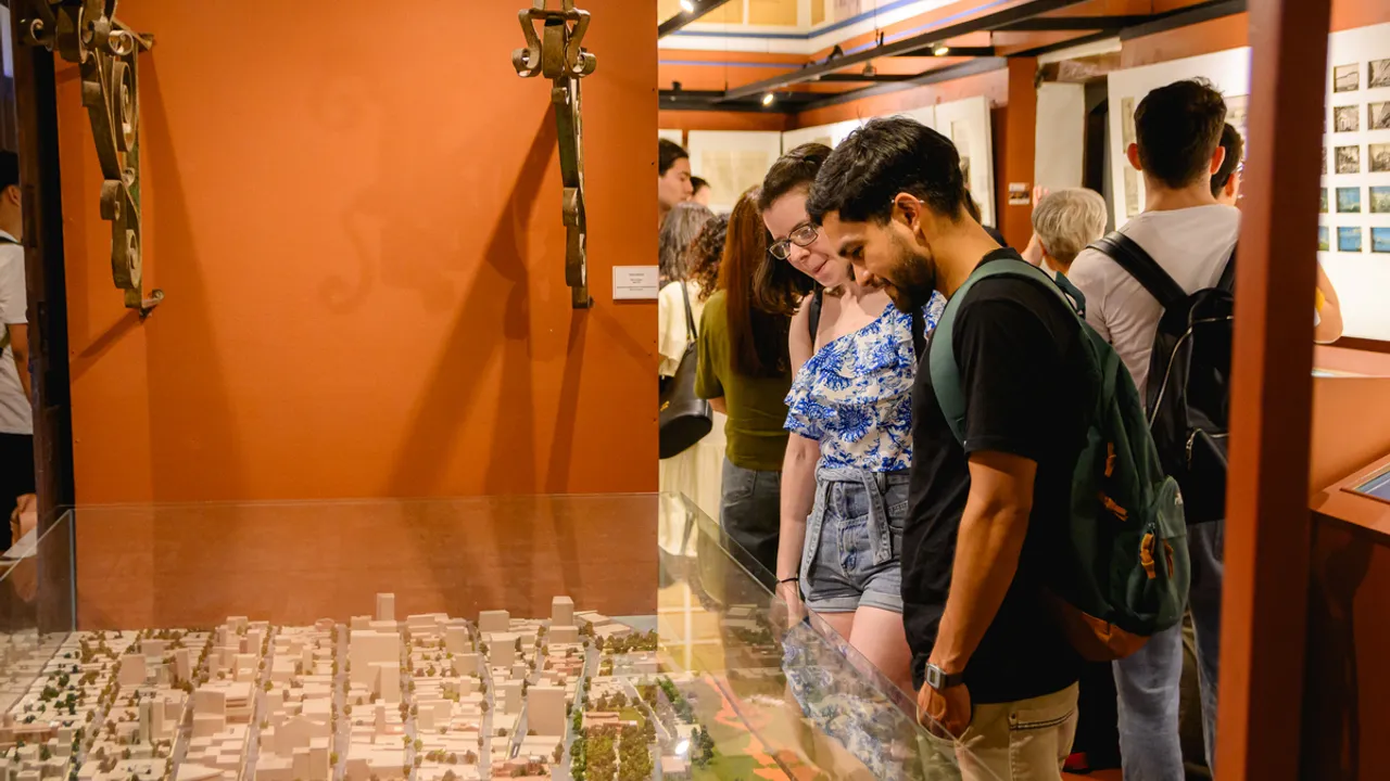 Dos jóvenes observan una maqueta urbana protegida por vidrio dentro de una sala de exposición cultural iluminada.